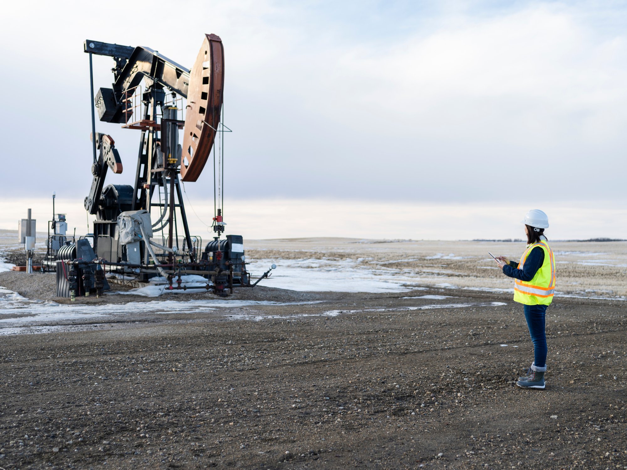 An oilfield worker wearing personal protective equipment holds a clipboard next to a pumpjack. 