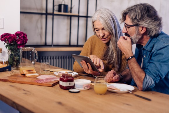 Two people look thoughtfully at tablet while sitting at table at home eating breakfast.
