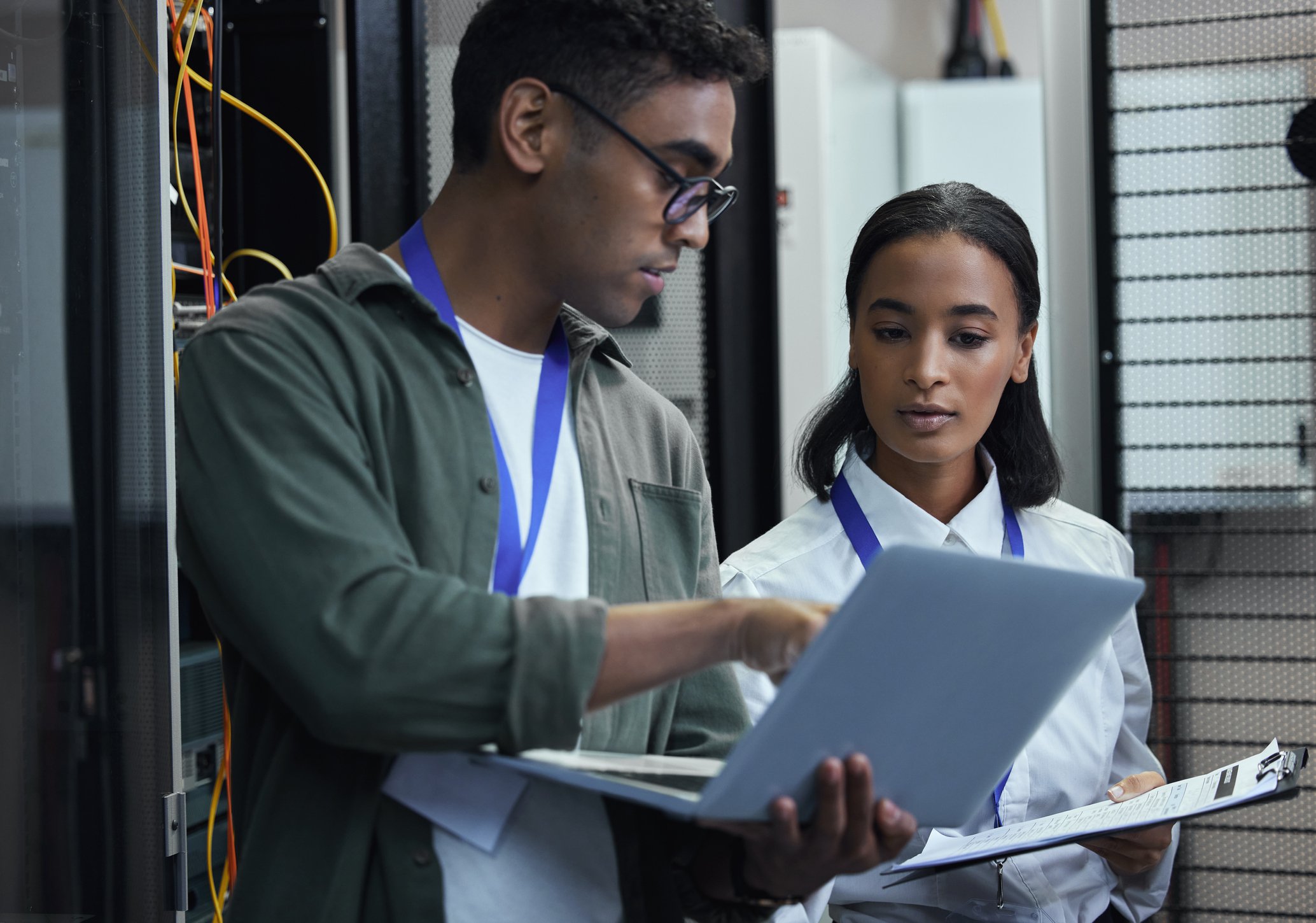 Two people in a server room. One is holding a laptop while the other holds a clipboard.