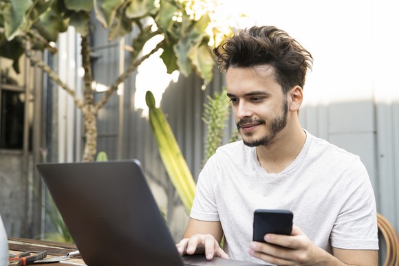 A person sits outside working on a laptop and holding a smartphone.