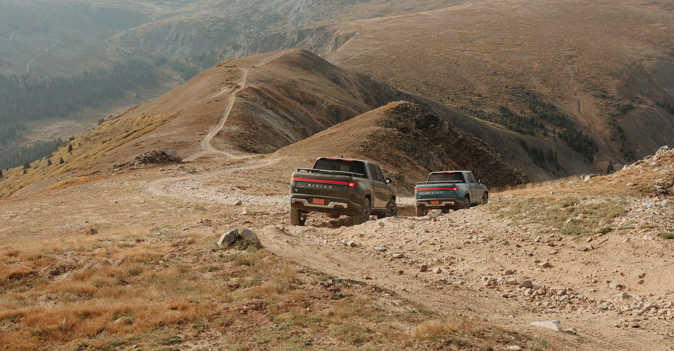 Rivian R1T pickup trucks driving away on mountain trail. 