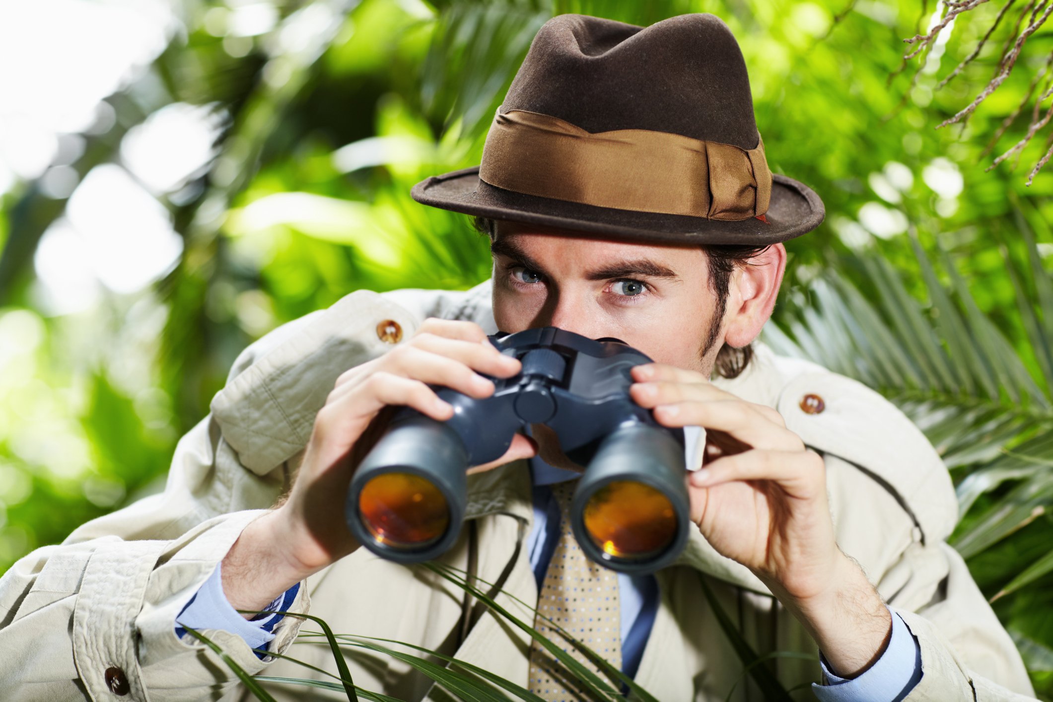 A detective holding binoculars hides in a bush.