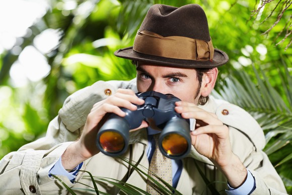 A detective holding binoculars hides in a bush.