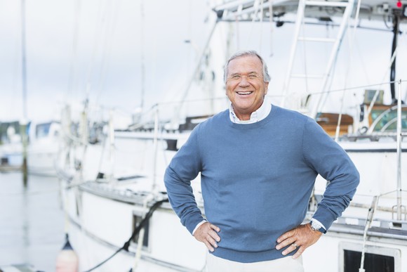 Affluent smiling man in a sweater standing on a dock in front of his yacht.