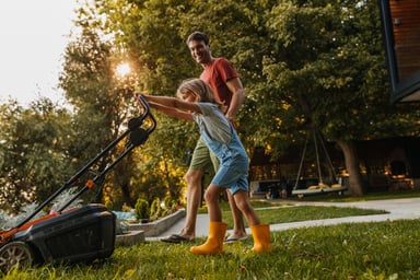 A parent helps a child mow the lawn.