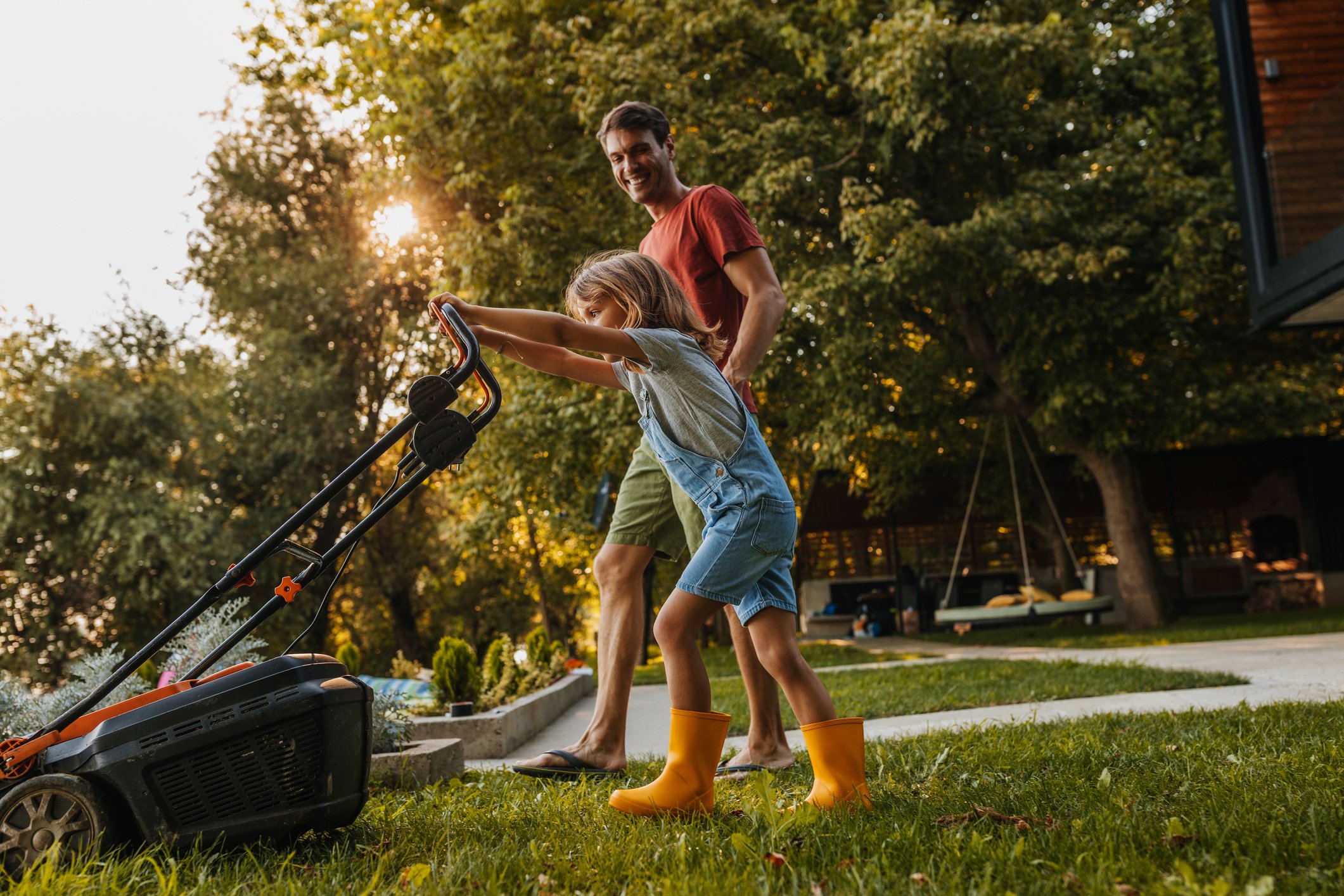 A parent helps a child mow the lawn.