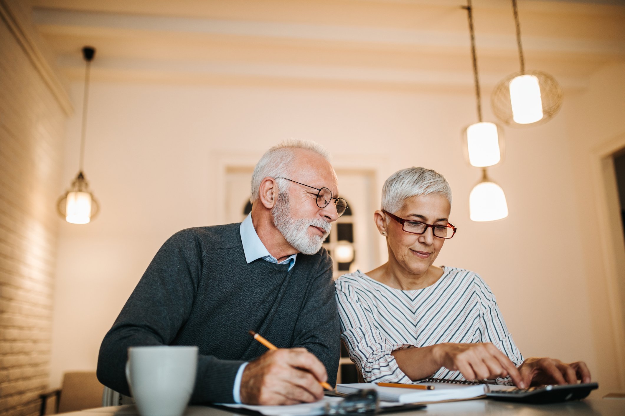 An older couple using a calculator and writing down notes at the kitchen table.