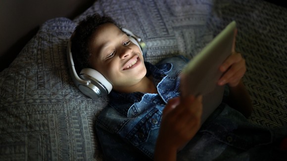 A young boy with headphones on, laying down, enjoying content on  a tablet.