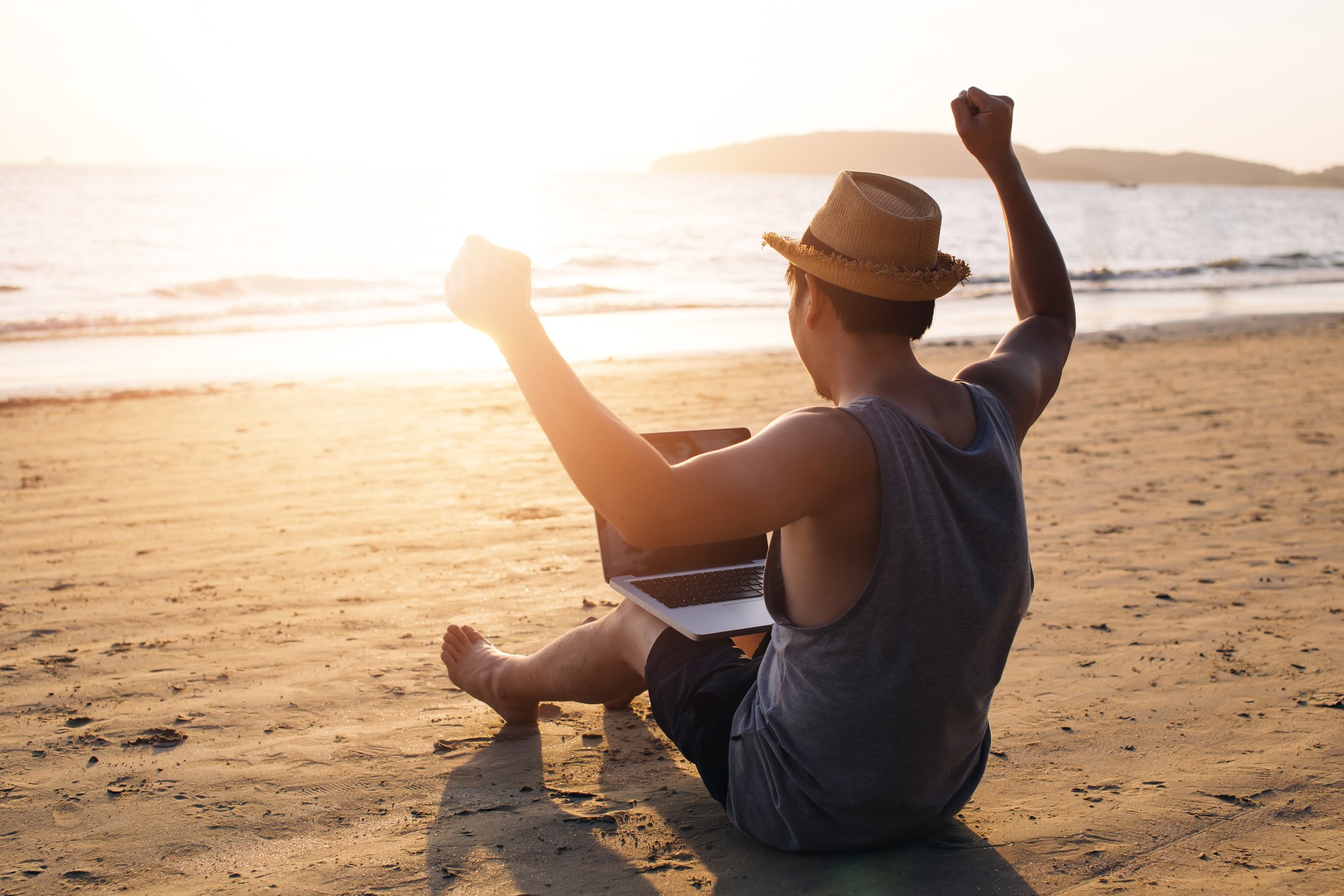 View from behind of person sitting on beach with laptop, arms raised in celebration.