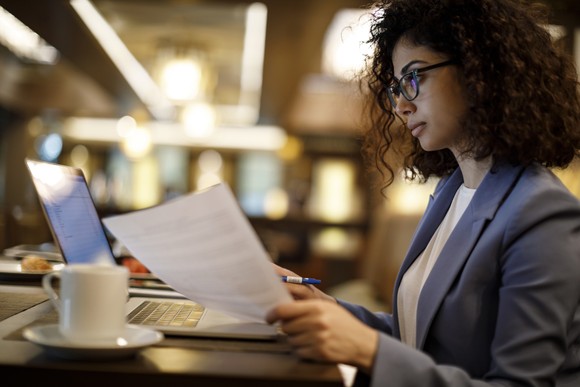 A person looking at a computer and holding a printout in a coffee shop.