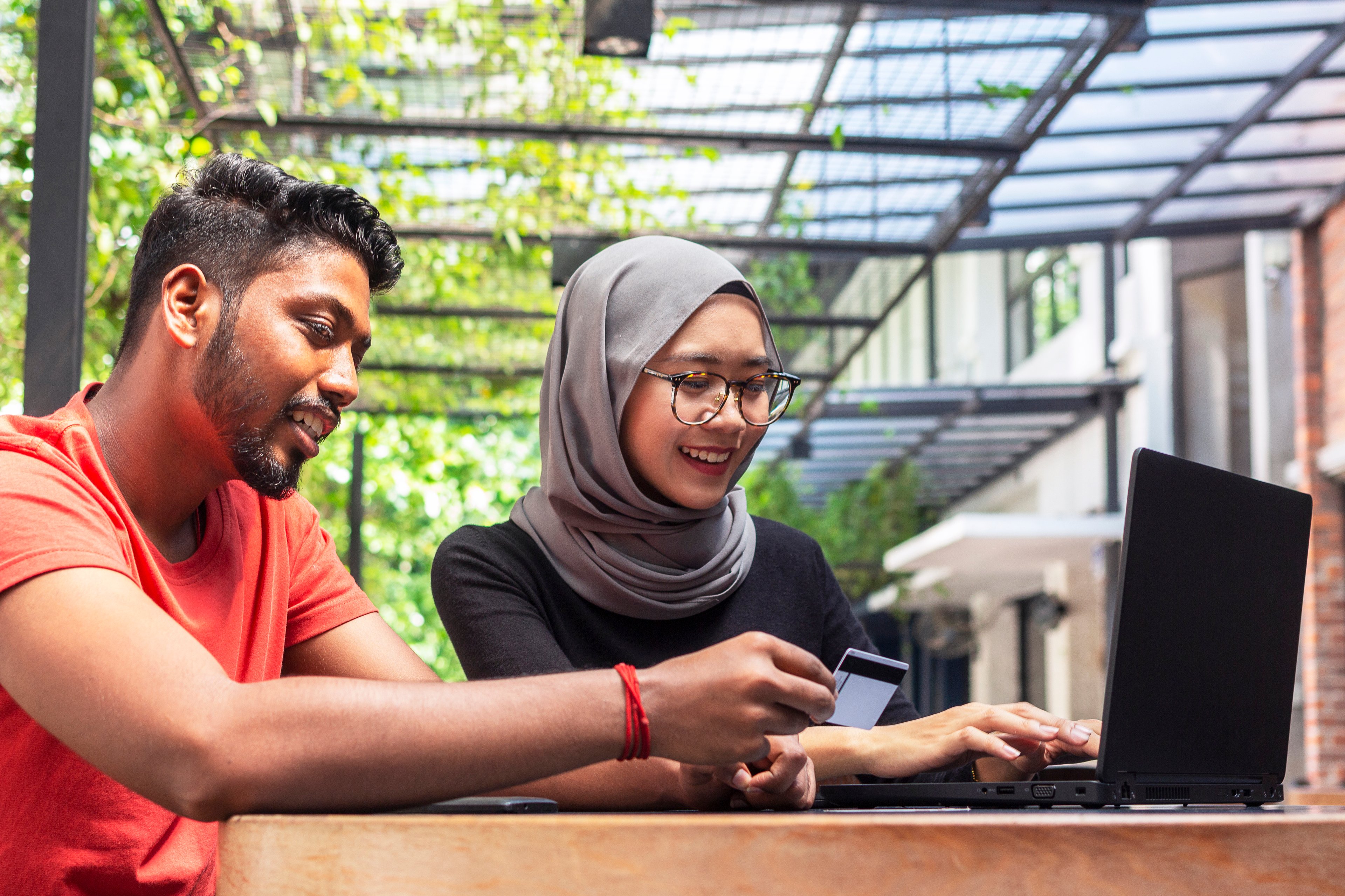 Person in orange t-shirt and person in hijab work together on laptop.
