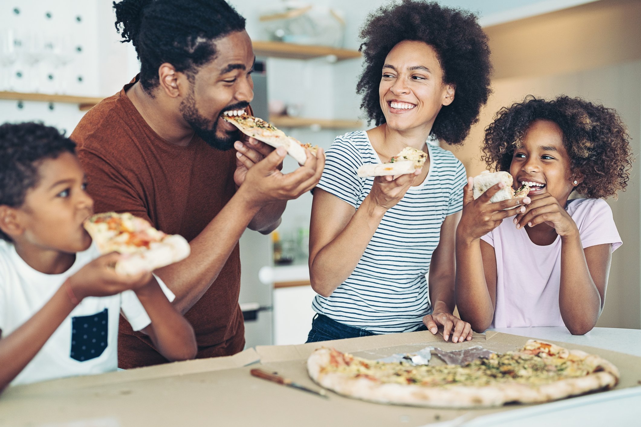 People smiling and eating pizza in a kitchen.