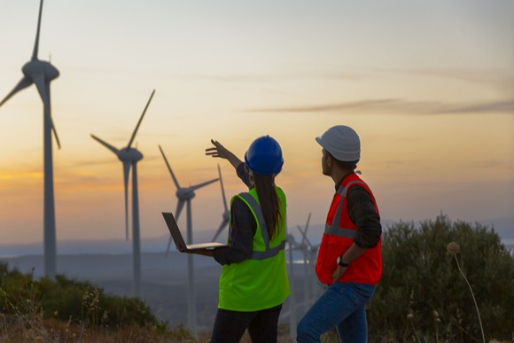 Two workers in hardhats and protective vests look at a row of wind turbines at sunset. 