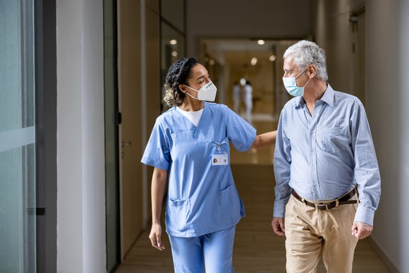 A healthcare worker in a blue uniform puts their arm behind an older adult.