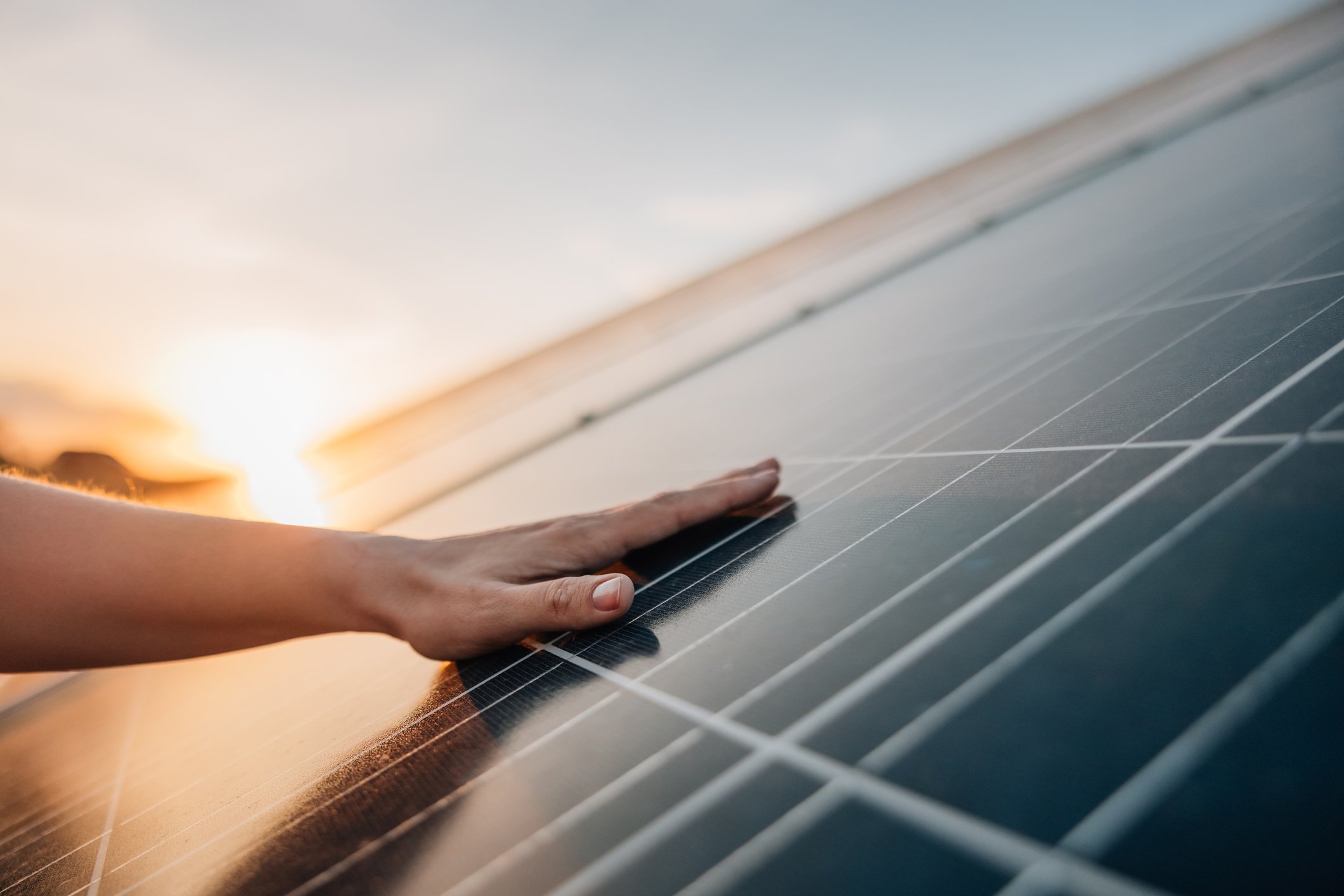 A person puts their hand on a solar panel as the bright yellow sun shines in the background. 