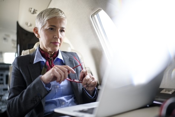 A woman sits in front of a laptop while in a jet. She is holding her pink glasses.
