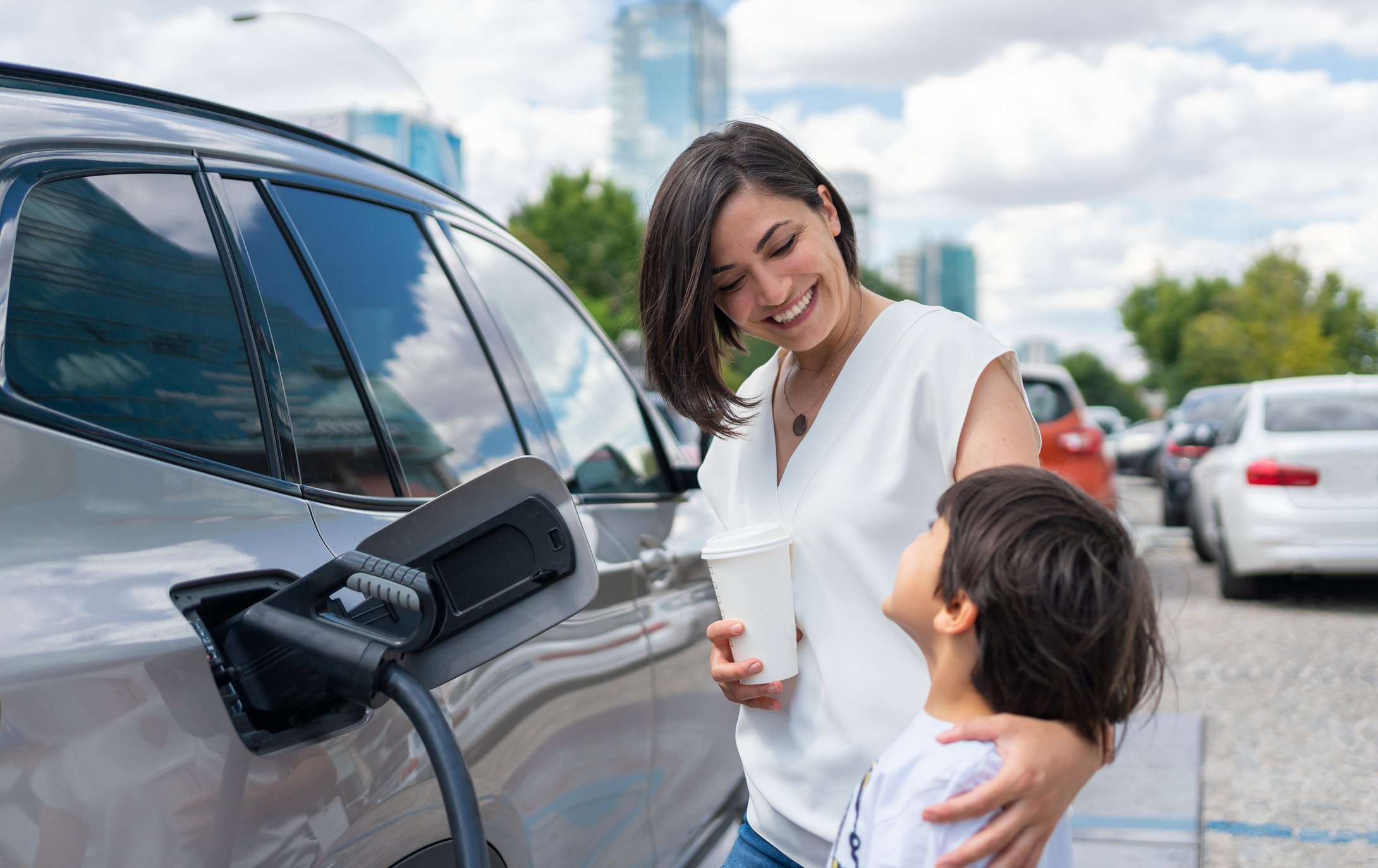 Two smiling people wait for an electric vehicle to charge at a charging station in a city.