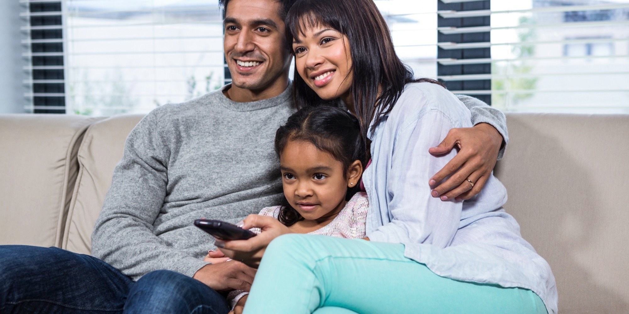 A couple and a child sit on a couch, watching TV.