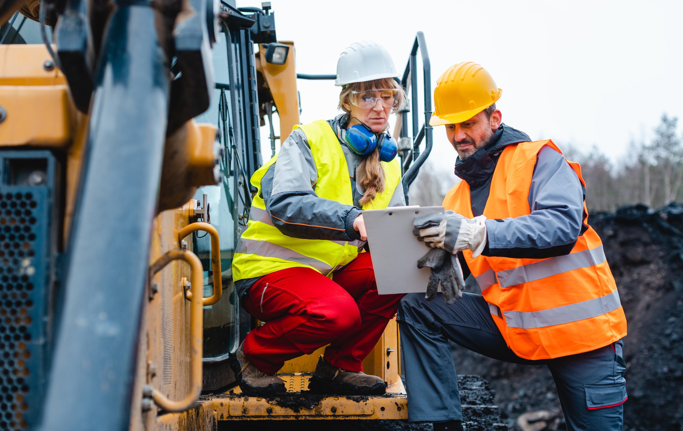 Two miners discuss a field report while one of them is sitting on a massive construction machine.