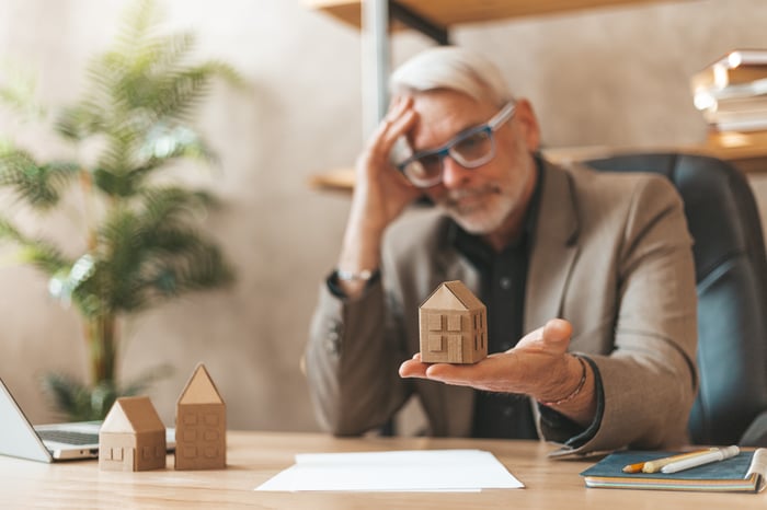 Person holding a block home in hand at desk while holding head.