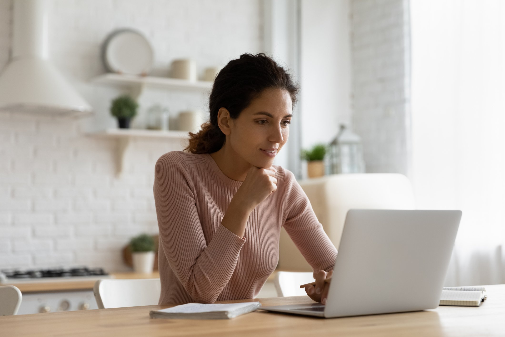 A person sitting at a kitchen table and reading something on a laptop.