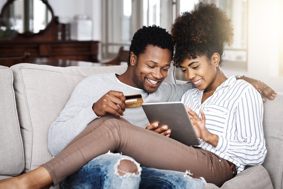 A couple sits on a couch looking at a tablet while holding a credit card.