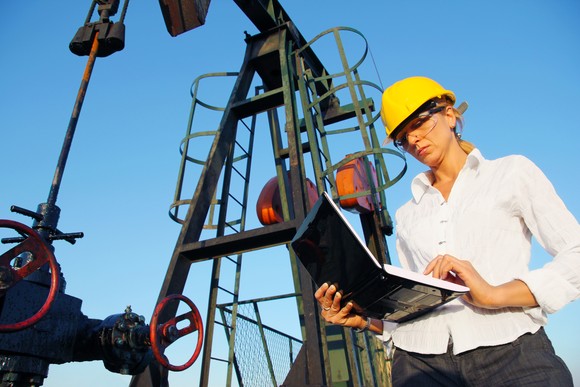 A person wearing a hard hat looking at a laptop while standing in front of an oil rig.