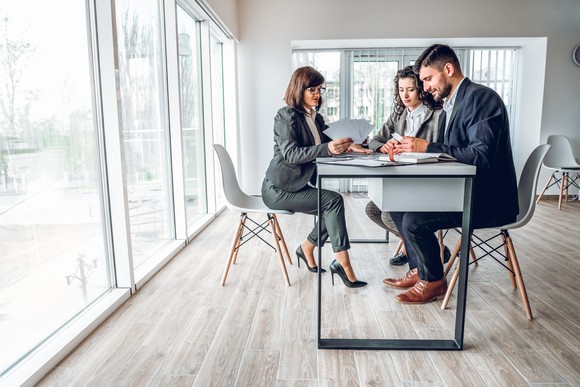 Three professionals review papers at a desk in an office.