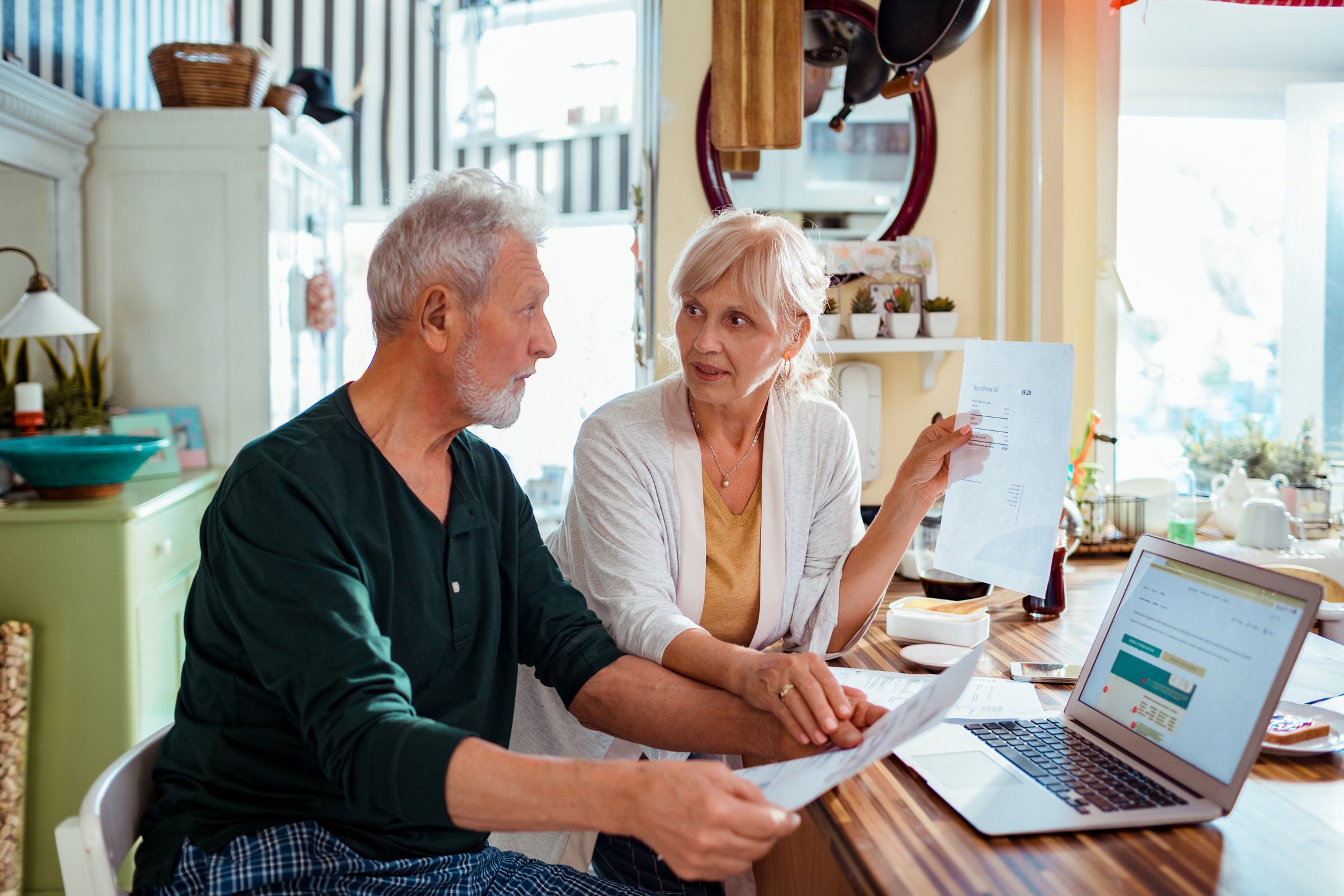 Two older adults talking and holding papers in front of a laptop.