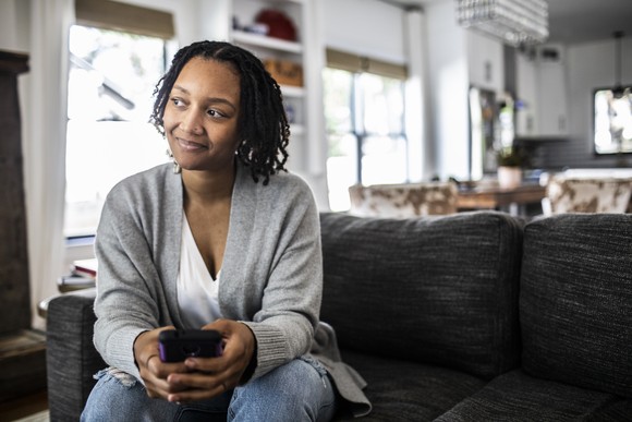 Person sitting on couch holding smartphone smiles thoughtfully.