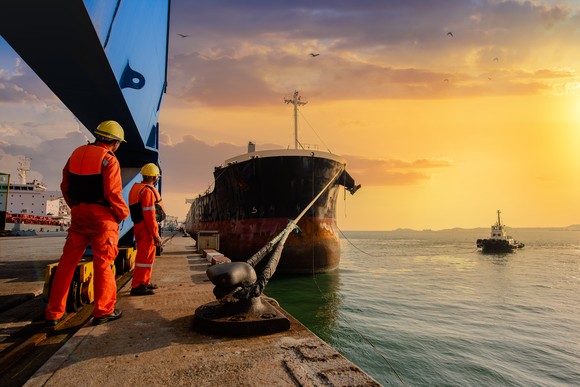 Dockworkers stand on a dock looking toward a freighter tied to the dock as a tugboat approaches