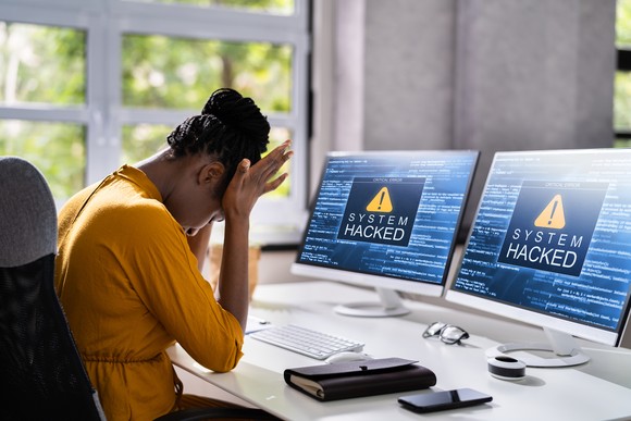 Upset person with their head in their hands, sitting at a desk, with computer monitors that read "system hacked".