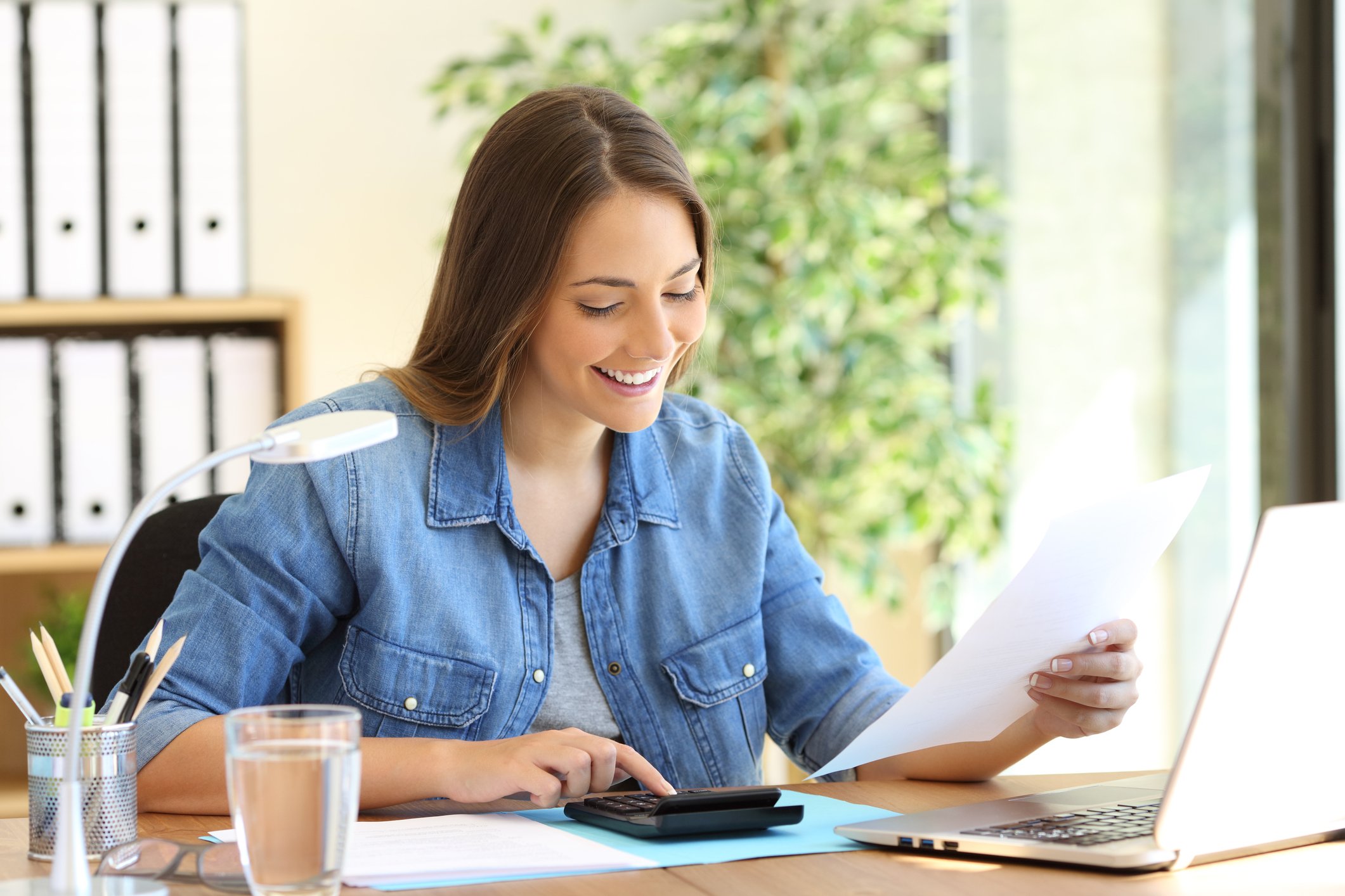 A person smiles while reading documents and using both a calculator and a laptop.