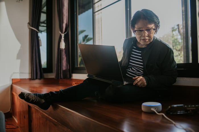 A person sits with a laptop and leans toward a phone.