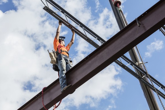 A worker wearing a hard hat, orange T-shirt, and jeans stands on a steel girder with a bright blue sky and sparse clouds in the background. 