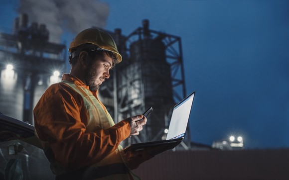 A refinery worker looks at a laptop at night in front of a petrochemical plant.