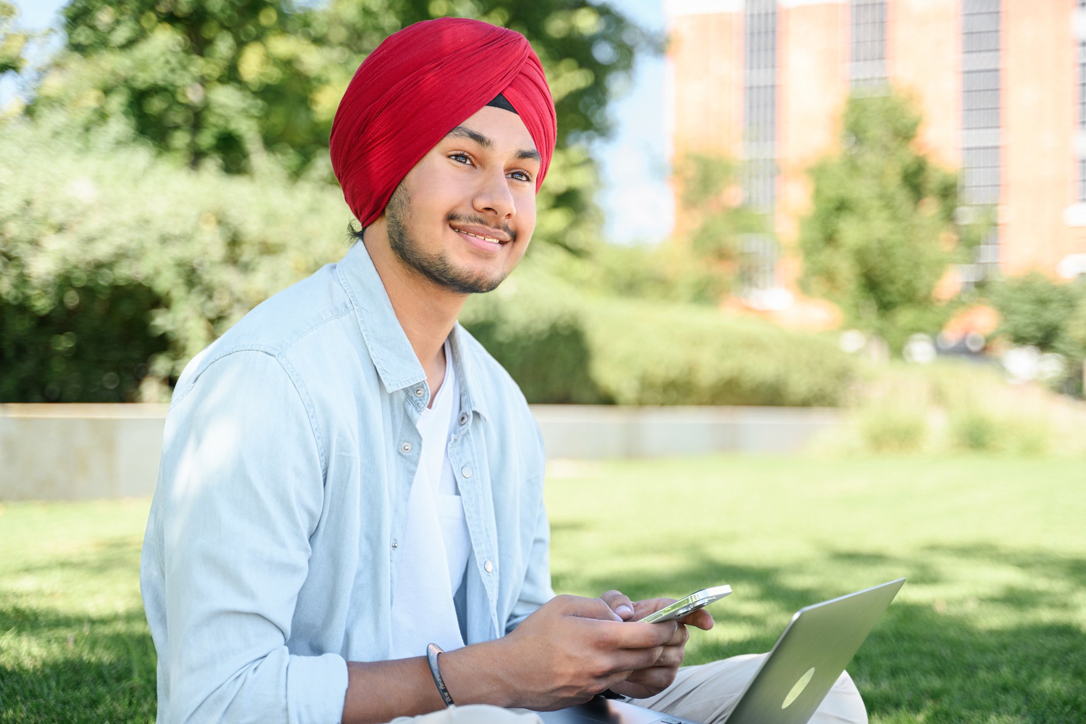 Someone is sitting outdoors, smiling with a phone and laptop.
