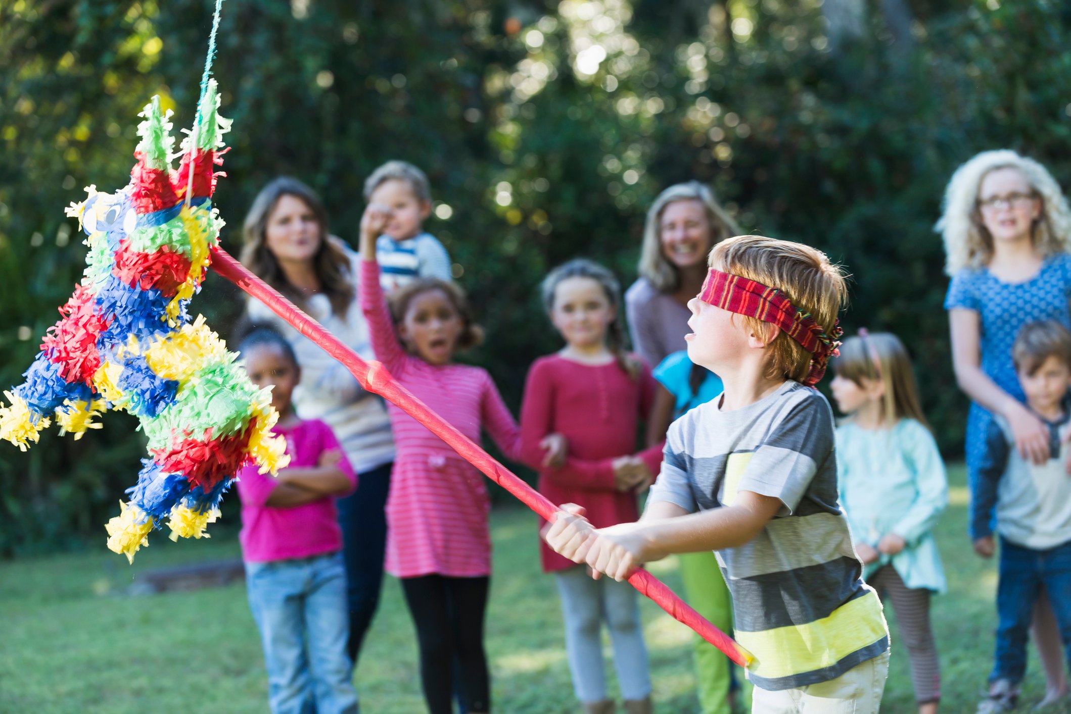 Blindfolded child hitting a pinata with a red stick while other children watch.