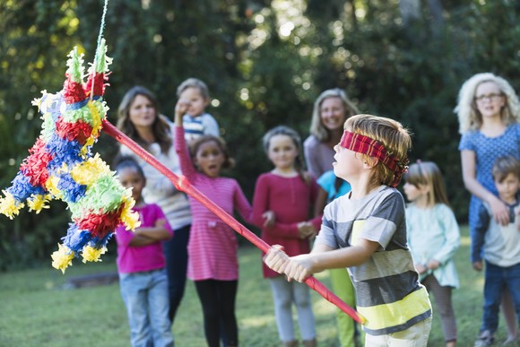 Blindfolded child hitting a pinata with a red stick while other children watch.