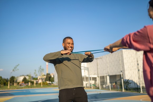 A person grimaces while doing a rubber band exercise with a partner outdoors. 
