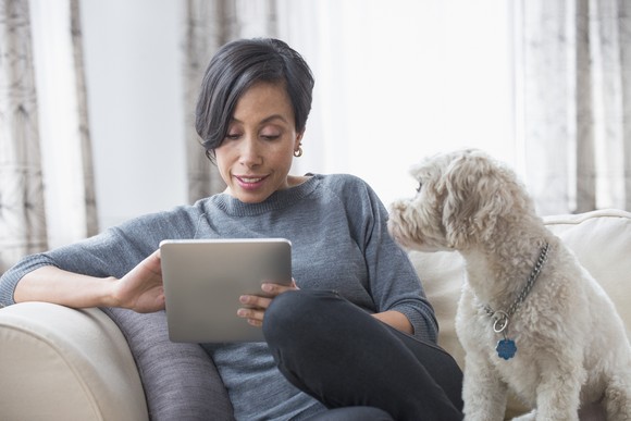 Person sitting on a couch with a dog while looking at a tablet.