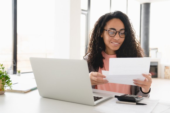 Person sitting at a table with a laptop and calculator in front of them while looking at a piece of paper.