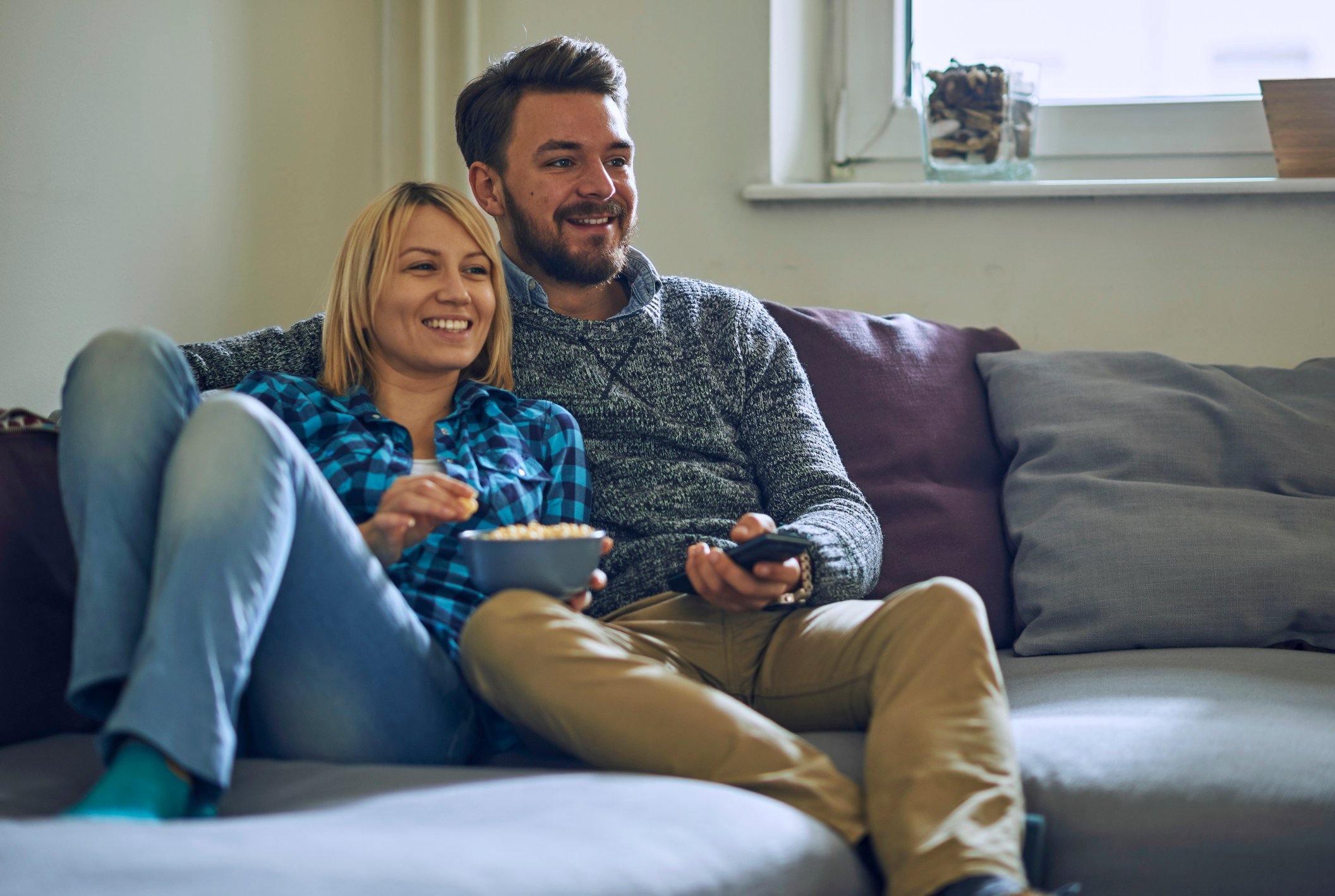 Two people holding a bowl of popcorn and a remote control while sitting on a couch.
