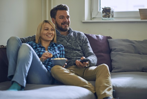 Two people holding a bowl of popcorn and a remote control while sitting on a couch.
