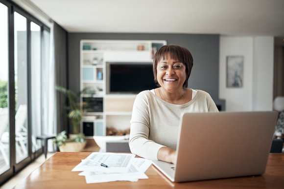 Person smiles while sitting in front of laptop at home with paperwork on the table. 