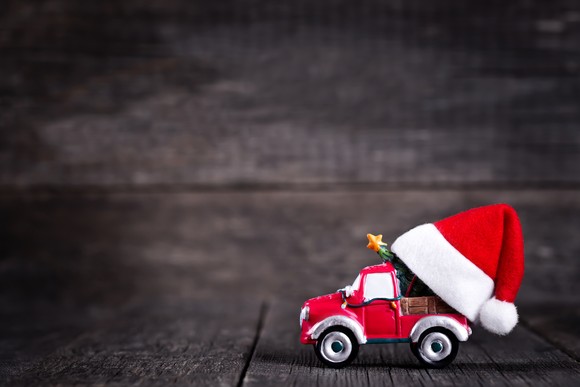 A red toy truck with a tree in the bed and a red Santa Claus hat sits in front of a wooden rustic background.