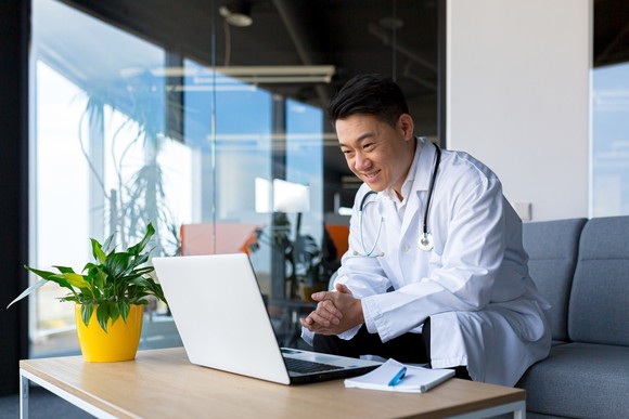 Doctor wearing a lab coat and stethoscope sitting on a coach at home, having a video conference on a laptop.