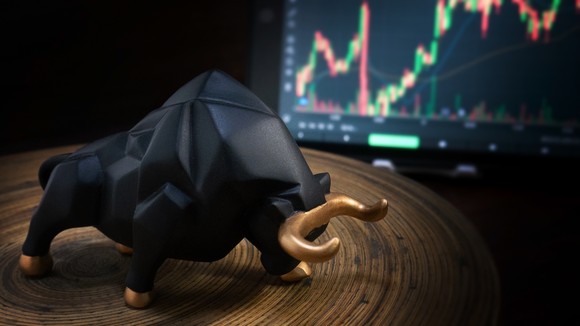 A bull figurine sits on a table in the foreground, while a monitor displays a stock price chart in the background.