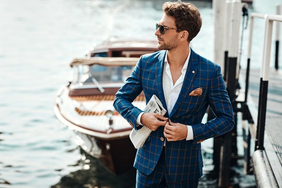 A person in a tailored suit stands on a dock in front of a boat. 