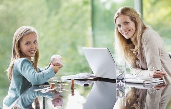 A child holding a piggy bank sits at a table with an adult who is using a laptop.